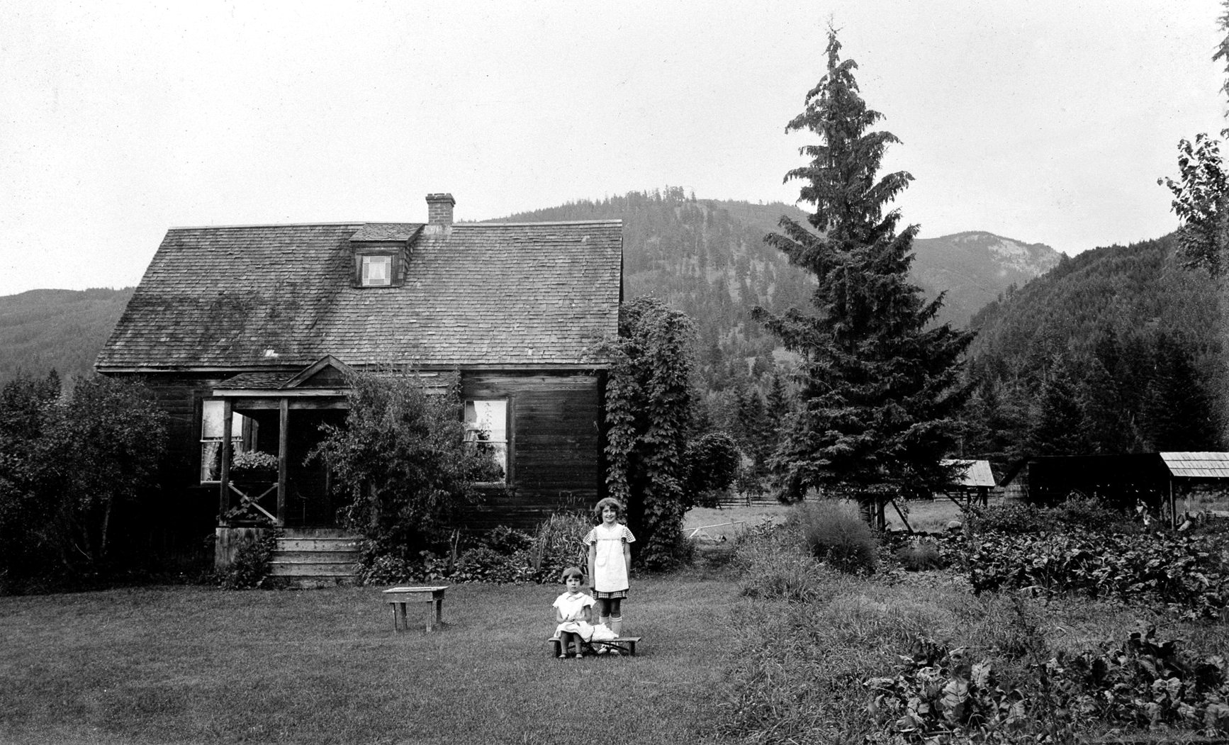 Two girls, Anne and Mary Meggait are posed in front of a small house on a homestead on Mountain View Road. The house has three windows of which one is in the peaked roof. A tall shrub is in front of the covered porch. Hills and trees surround the homestead . Two small sheds are in the background. This is the original house and it burnt down according to notes with picture envelope
